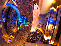 Close-up of a glass lens being processed in a machine with coolant and sparks.