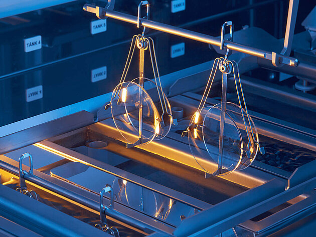 Semiconductor wafers suspended by metal holders above a processing bath in a cleanroom environment.