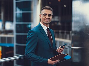 Businessman in a suit holding a smartphone and a notebook, standing in a modern office building.