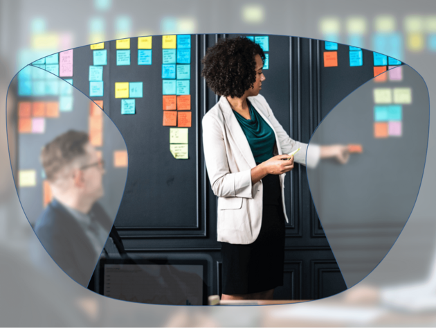 View through a pair of glasses showing a woman presenting at a board covered with colorful sticky notes, while colleagues listen.