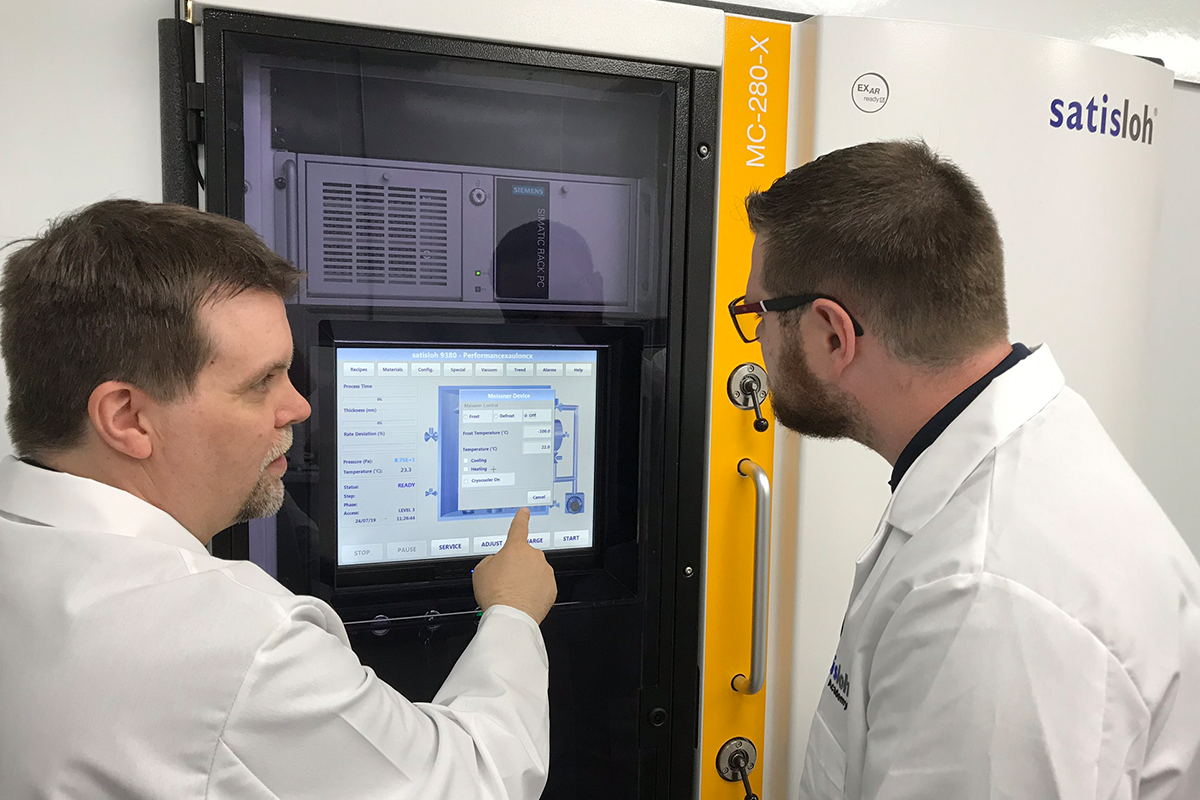 Two technicians in lab coats operating a machine using a touchscreen control panel.