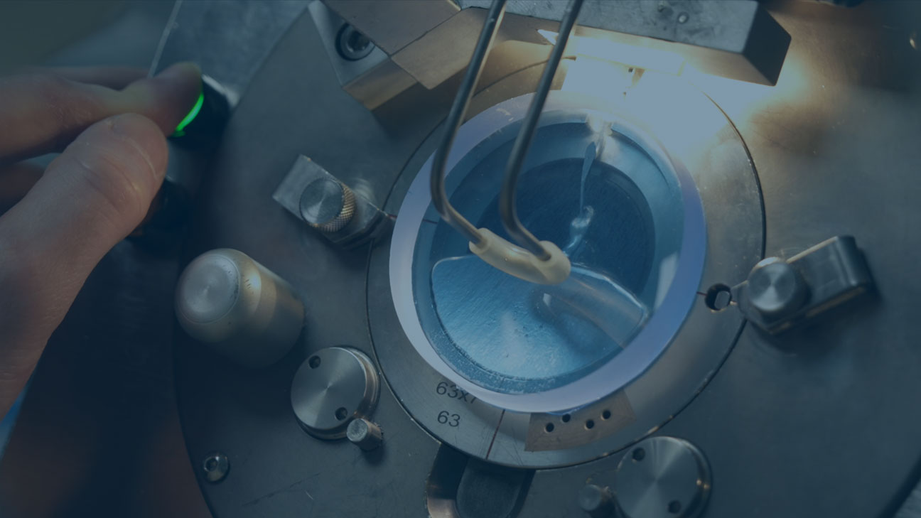 Technician’s hand operating a machine that precisely processes a glass lens with fine tools