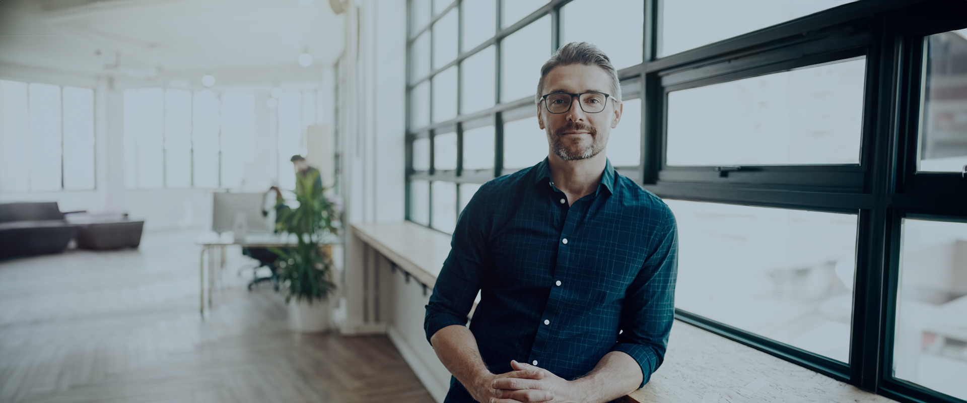 Genießen Sie nahtlose Klarheit Man with glasses and a beard sitting by a large window in a bright, modern office space.