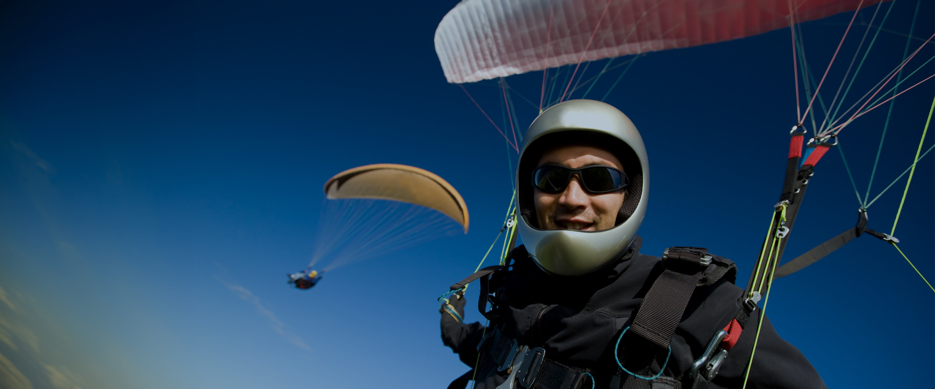 Aktiven Lebensstil verbessern Paraglider wearing a helmet and sunglasses smiling at the camera, with another paraglider visible in the background against a clear blue sky.
